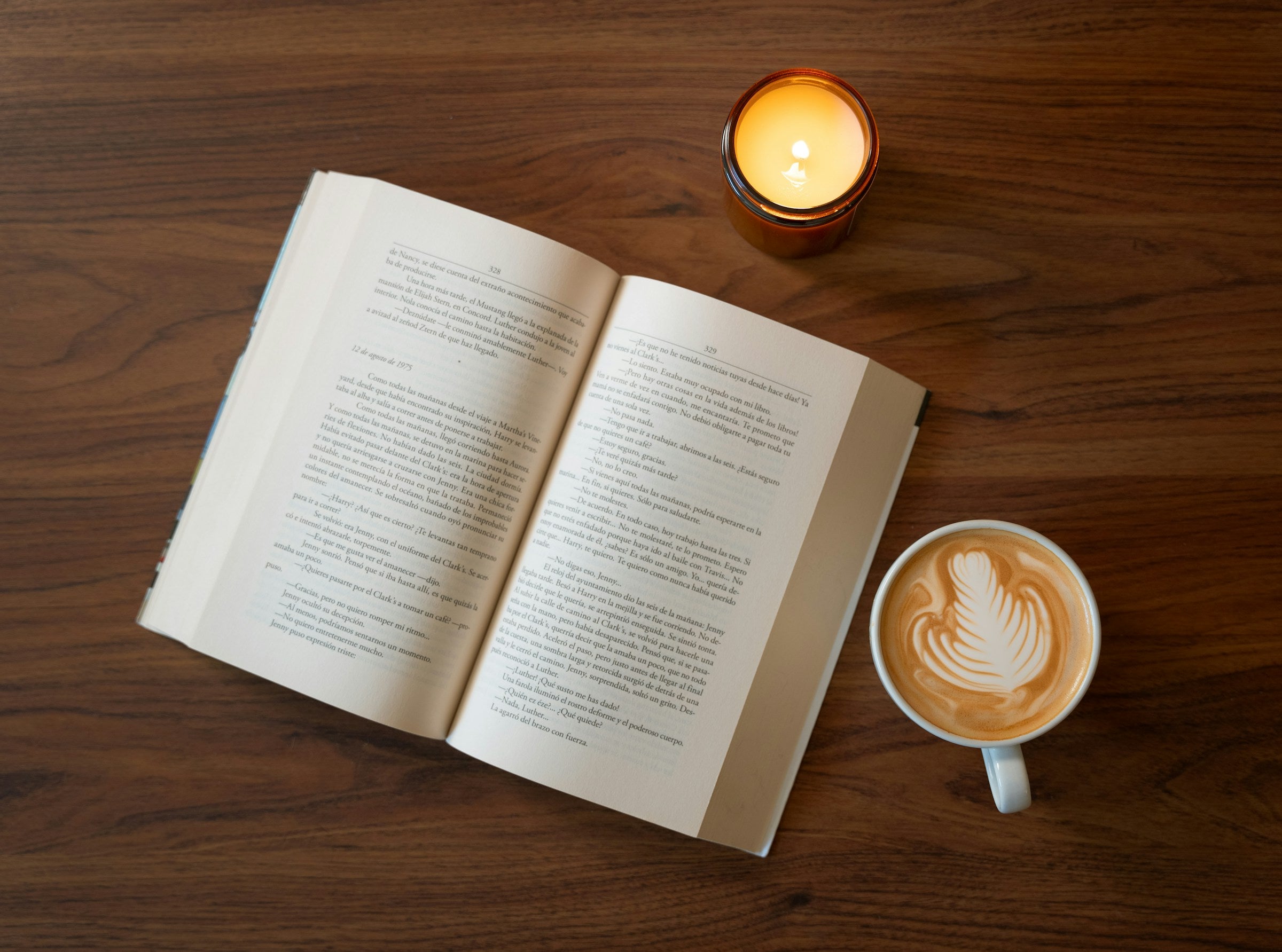 Open book with latte art coffee and lit candle on wooden table for cozy reading